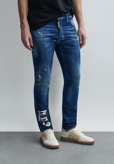 Jean en denim bleu à coupe slim, avec des éclaboussures de peinture et des accents graphiques colorés sur la jambe gauche. Associé à des baskets beiges.