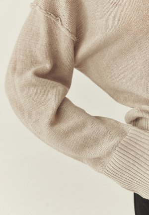 Ærme af beige strikket sweater med ribkant og en diskret sømdetalje nær skulderen, vist på en bøjet arm mod en lys baggrund.