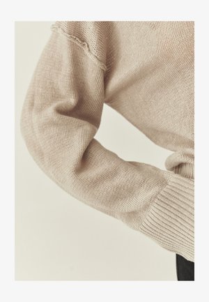 Ærme af beige strikket sweater med ribkant og en diskret sømdetalje nær skulderen, vist på en bøjet arm mod en lys baggrund.