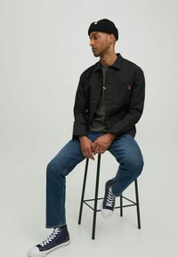 Sort skjorte med knapper, grøn t-shirt, blå jeans og marineblå sneakers. Tilbehøret inkluderer en sort strikket beanie. Model sidder på en sort skammel.