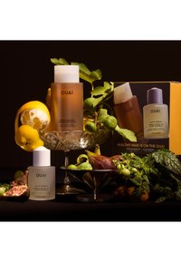 Bottiglie di shampoo e siero per il cuoio capelluto OUAI in vetro dorato e opaco, esposte con frutta assortita e verdure su uno sfondo scuro.