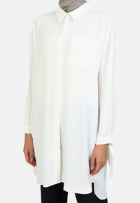 Camicia bianca lunga, realizzata in tessuto leggero, con colletto punta, taschino sul davanti e maniche aperte con accentuazioni a nastro.