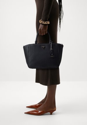Femme portant une robe marron côtelée tenant un sac à main noir tressé de la marque Guess, chaussures en cuir verni marron à talons bas pointus et un bracelet chaîne en or.