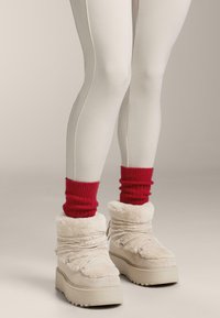 Enkellaarsjes van nepbont in beige met een dikke witte zool, voorzien van gekruiste bandjes en zachte rode gebreide beenwarmers.