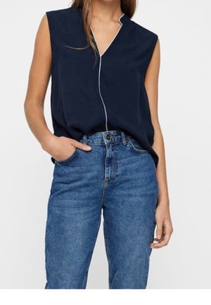 Vrouw draagt een mouwloze marineblauwe blouse met witte bies en een hoge taille blauwe spijkerbroek, hand in de zak tegen een effen achtergrond.