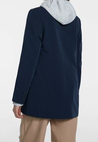 Blazer navy con cappuccio grigio, caratterizzato da un design su misura, tessuto morbido e polsini a quattro bottoni sulle maniche.