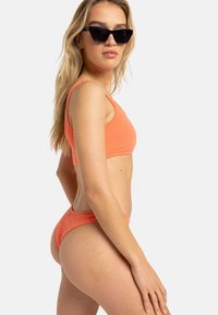 Bikini texturé orange vif avec un haut ajusté et un bas assorti. La mannequin porte des lunettes de soleil noires angulaires, posant de profil.