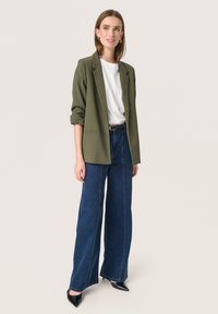 Olivově zelený blazer přes bílou košili, spárovaný s širokými modrými denímovými kalhotami a černými špičatými lodičkami. Jednoduchý design, strukturovaný střih.
