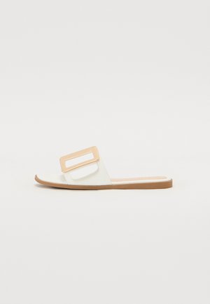 Sandale à glissière avec une partie supérieure blanche à finition texturée et un accent rectangulaire doré. Une semelle plate brune complète le design.
