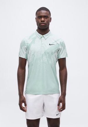 Mann steht, trägt hellgrünes, gemustertes Polo und weiße Shorts mit schwarzen Nike-Logos auf einfarbigem weißem Hintergrund.
