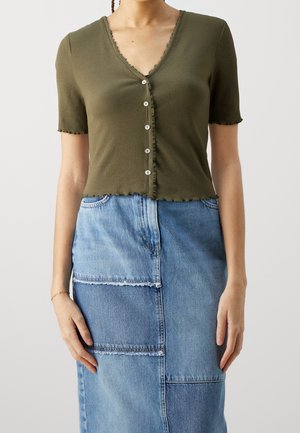 oliwkowy, żebrowany top z krótkim rękawem, z dekoltem w serek, z przodu na guziki i falowanym dołem. W zestawie z jasnoniebieską spódnicą z denimu z łatami i postrzępionymi brzegami.