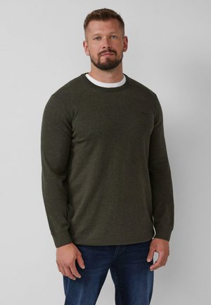 Mann mit leichtem Bart, der einen dunkelgrünen Langarmpullover über einem weißen Hemd und blauen Jeans trägt, steht vor einem einfarbigen hellen Hintergrund.