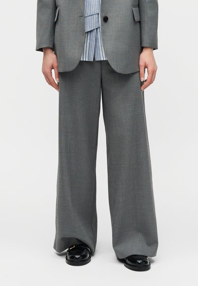 Personne portant un pantalon large gris, un blazer assorti, une chemise à rayures bleues et des mocassins noirs, debout devant un fond blanc.