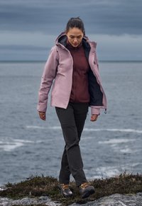 Woman in outdoor gear walking on rocky ground near ocean under cloudy sky, wearing a pink jacket, maroon top, black pants, and hiking boots.