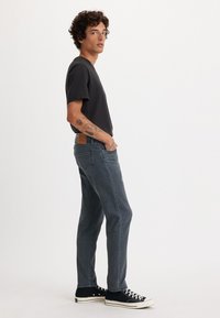 Czarna koszulka, ciemnoniebieskie dżinsy i czarne trampki typu high-top. Dżinsy mają brązową łatę z tyłu i lekko wyblakłe miejsca. Model stoi bokiem.