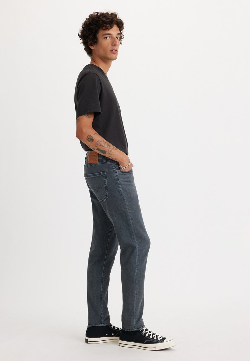 Schwarzes T-Shirt, dunkelblaue Jeans und schwarze High-Top-Sneaker. Die Jeans haben einen braunen Fleck auf der Rückseite und eine leichte Verblasserung. Das Model steht seitlich.