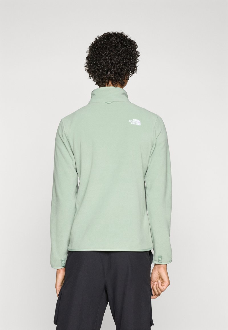 Grön fleece-pullover med hög krage och en synlig vit logotyp på den övre ryggen. Har långa ärmar och en figurnära design.