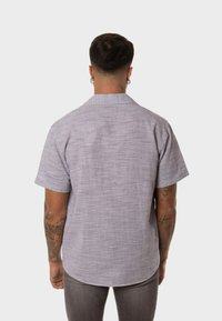 Camisa de manga corta en color púrpura claro, con tejido texturizado y cuello. La vista posterior muestra un dobladillo curvado y un ajuste relajado.