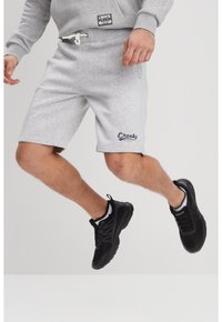 Pantalones cortos de algodón grises con bolsillos laterales y cintura ajustable con cordón, que presentan un logo azul de "Creeks". Llevados con zapatillas deportivas negras.