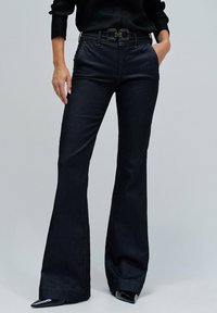 Los pantalones acampanados de mezclilla oscura presentan una cintura alta con un cinturón negro de doble hebilla, bolsillos laterales y ligeros detalles de costura en las costuras.
