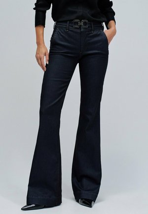Los pantalones acampanados de mezclilla oscura presentan una cintura alta con un cinturón negro de doble hebilla, bolsillos laterales y ligeros detalles de costura en las costuras.