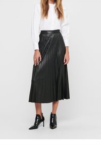 Jupe midi plissée noire en simili cuir, taille haute avec une finition brillante, assortie à une blouse blanche à manches longues et des bottines noires.