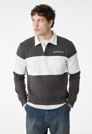 Junger Mann, der nach links schaut, trägt ein dunkelgraues und weißes Rugby-Shirt mit gesticktem Text und blaue Jeans, Hände in den Taschen.