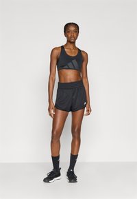 Μαύρο αθλητικό σουτιέν με υφή ύφανσης και λογότυπο, σε συνδυασμό με μαύρα σορτς με ελαστική μέση και λογότυπο. Μαύρες κάλτσες και αθλητικά παπούτσια.