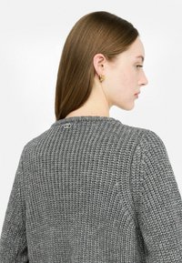 Maglione grigio lavorato a maglia con un motivo texturizzato, scollatura tonda e un piccolo logo sulla schiena. Chi lo indossa ha capelli lunghi e orecchini d'oro.