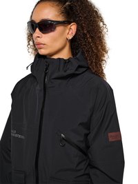 Frau mit lockigem Haar, die schwarze Wraparound-Sonnenbrille und eine schwarze Kapuzenjacke mit Reißverschlusstaschen und einem roten Aufnäher am Ärmel trägt.