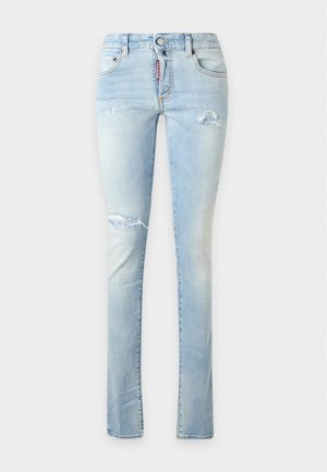 Femme debout, une main sur la hanche, portant un haut marron moulant à manches courtes, un jean skinny bleu clair déchiré et des sandales noires à talons.