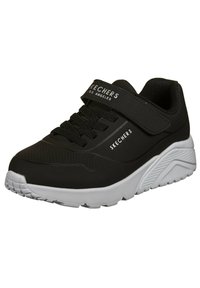 Sneaker nera con lacci e cinturino in velcro, realizzata in materiale sintetico. Presenta una suola bianca e dettagli testurizzati sui lati.