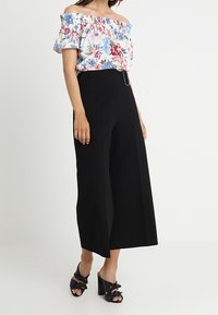 Donna con una blusa floreale a spalle scoperte, pantaloni cropped neri a vita alta e gamba larga e sandali neri con tacco e fiocchi.