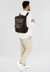 Zaino in pelle marrone con molteplici tasche, texture liscia e cerniere argentate. Indossato con pantaloni chiari e sneakers bianche.