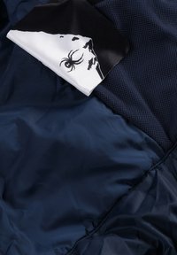 Marineblaue Jacke aus glattem, glänzendem Stoff mit Mesh-Details. Ein angenähtes Etikett zeigt eine schwarz-weiß Spider-Grafik.