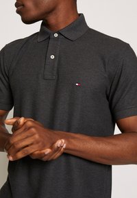 Polo gris avec une patte de boutonnage à trois boutons, col classique, et un petit logo brodé en rouge, blanc et bleu sur la poitrine.