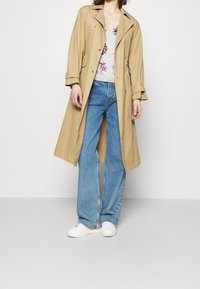 Beige trench coat with a belted waist, double-breasted design, and flap pockets, paired with light blue flared jeans and white shoes.