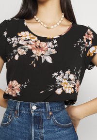 Top negro con flores de manga corta que presenta flores en tonos durazno y blanco, combinado con unos pantalones vaqueros de cintura alta azul. Adornado con un collar de conchas.