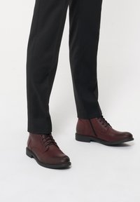 Burgundowe skórzane botki za kostkę z okrągłym noskiem, sznurowane z przodu, na czarnej gumowej podeszwie, zestawione z czarnymi dopasowanymi spodniami.