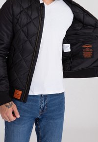 Veste bomber matelassée noire, dotée d'une fermeture éclair dorée et d'une étiquette orange sur l'ourlet inférieur. Poche intérieure et étiquette visibles à l'intérieur.
