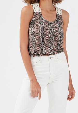 Femme portant un top sans manches à motifs avec des bretelles blanches en crochet et un jean blanc taille haute, debout les mains détendues le long du corps.