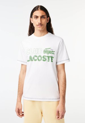 T-shirt blanc à manches courtes avec le texte "CLUB LACOSTE" en vert, arborant un logo de crocodile. Texture lisse, col rond, couture contrastante.