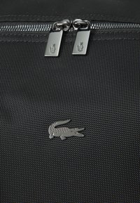 Čierna textúrovaná cestovná taška so zdvojeným zipsom so striebornými ťahadlami a metalickým logom krokodíla.