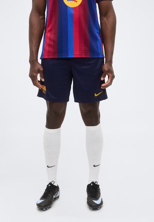 Homme portant un maillot de sport rayé bleu et rouge, un short bleu marine avec les logos du FC Barcelone et de Nike, des chaussettes blanches et des chaussures à crampons noires, debout sur fond blanc.