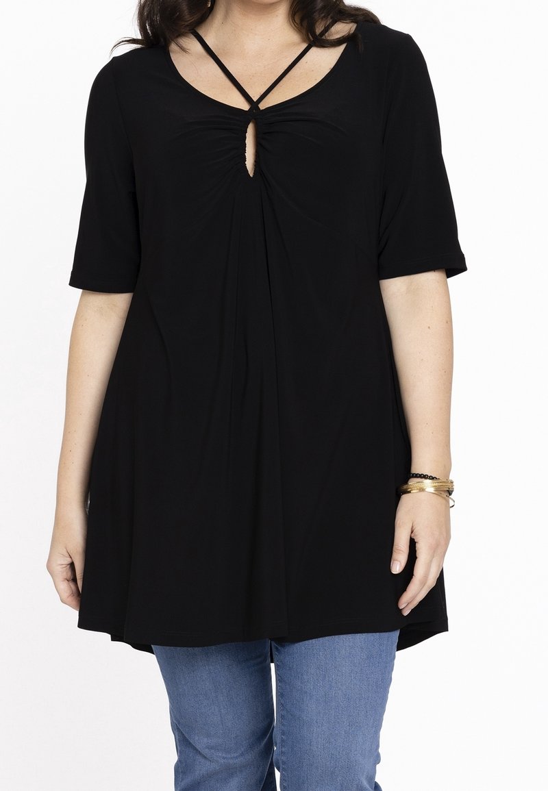 Femme portant un haut noir ample avec des bretelles croisées et un décolleté en trou de serrure, associé à un jean bleu et des bracelets dorés.