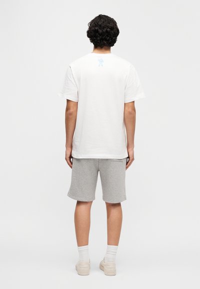 Λευκό βαμβακερό κοντομάνικο με ανοιχτό μπλε γραφικό στο πίσω μέρος, συνδυασμένο με γκρι σορτς και κρεμ αθλητικά παπούτσια, από πίσω όψη.