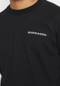 Sudadera negra con cuello redondo acanalado y mangas raglán. Presenta el logotipo en blanco "SCOTCH & SODA" en el área del pecho izquierdo. Textura suave.