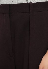 Burgunderfarbene maßgeschneiderte Hosen aus strukturiertem Stoff, mit gefaltetem Vorderteil, Gürtelschlaufen und Seitentaschen. Weiche Haptik und strukturierte Form.