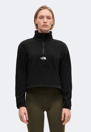 Personne aux cheveux blonds courts portant une polaire noire à fermeture éclair quart de zip avec le logo The North Face et un pantalon vert olive foncé, debout devant un fond uni.