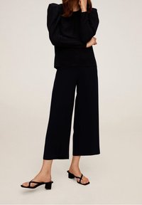Černý dlouhý rukáv top s vycpanými rameny, spárovaný s černými širokými kalhotami a černými nazouvacími sandály na podpatku. Hladká látka, minimalistický design.